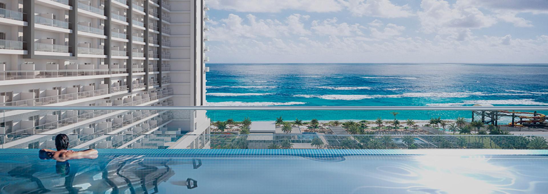 A person relaxing in an infinity pool with a panoramic view of the turquoise ocean and waves. A modern multi-story resort building with balconies is visible on the left, and palm trees line the beach in the distance.