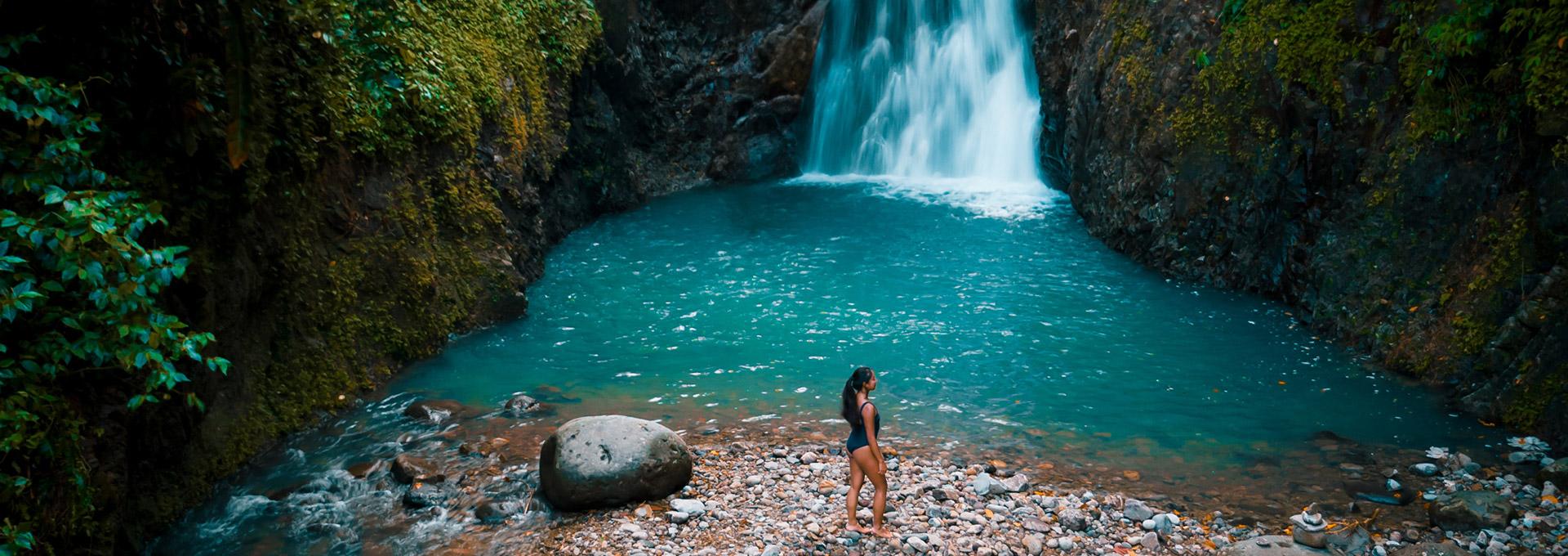 A person in a dark swimsuit stands on a rocky shore looking towards a stunning blue waterfall plunging into a clear turquoise pool, surrounded by dense green jungle foliage.