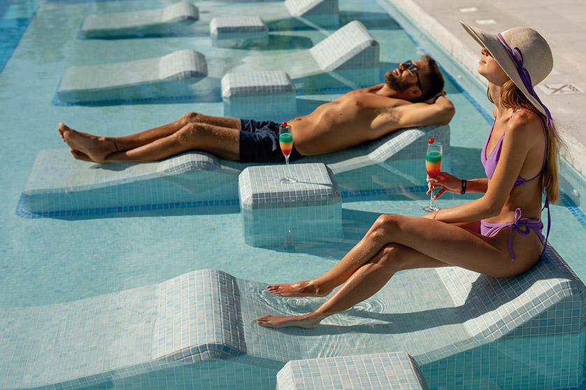 Idyllic pool scene with a couple enjoying refreshing drinks on submerged lounge chairs, evoking a sense of relaxation and vacation.