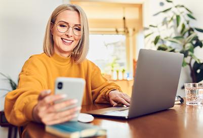 A happy woman engaging with her phone while working on a laptop, possibly in a home office setting.