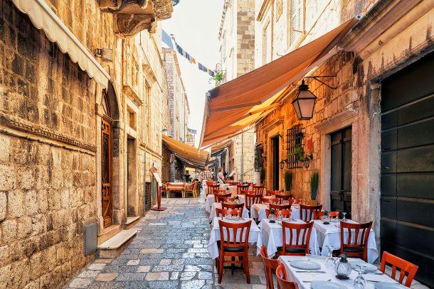 A charming outdoor restaurant with red chairs and white tablecloths set up on a cobblestone street, flanked by tall stone buildings with awnings in a historic European town.