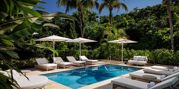 Tranquil resort pool scene featuring a clear blue pool, shaded lounge chairs with white cushions, and lush tropical greenery including palm trees.